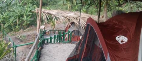 Een tent met een logo, een groen hek en een constructie met rieten dak in een natuurlijke omgeving.