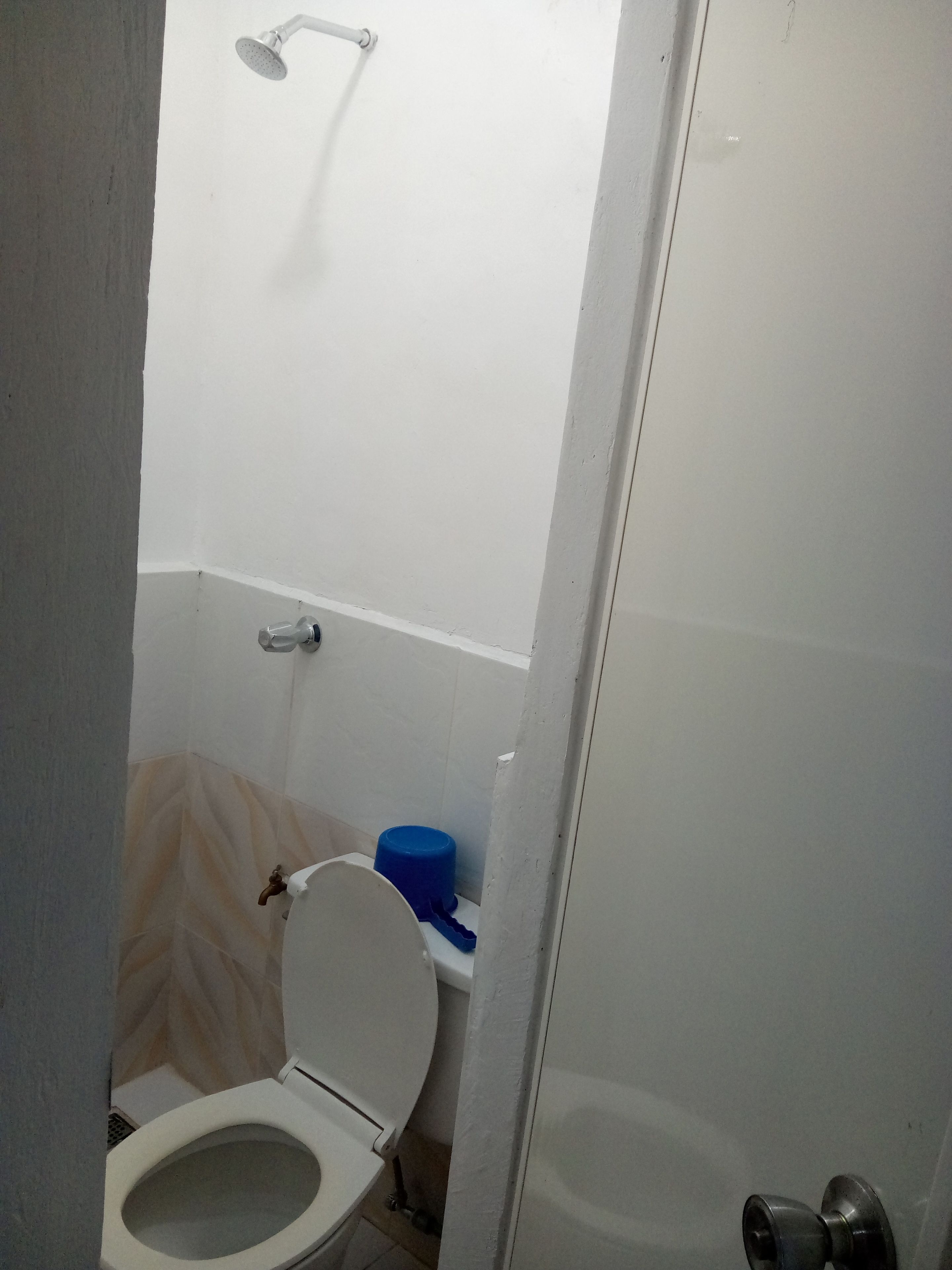 Um banheiro pequeno com vaso sanitário, chuveiro e um recipiente azul no chão.