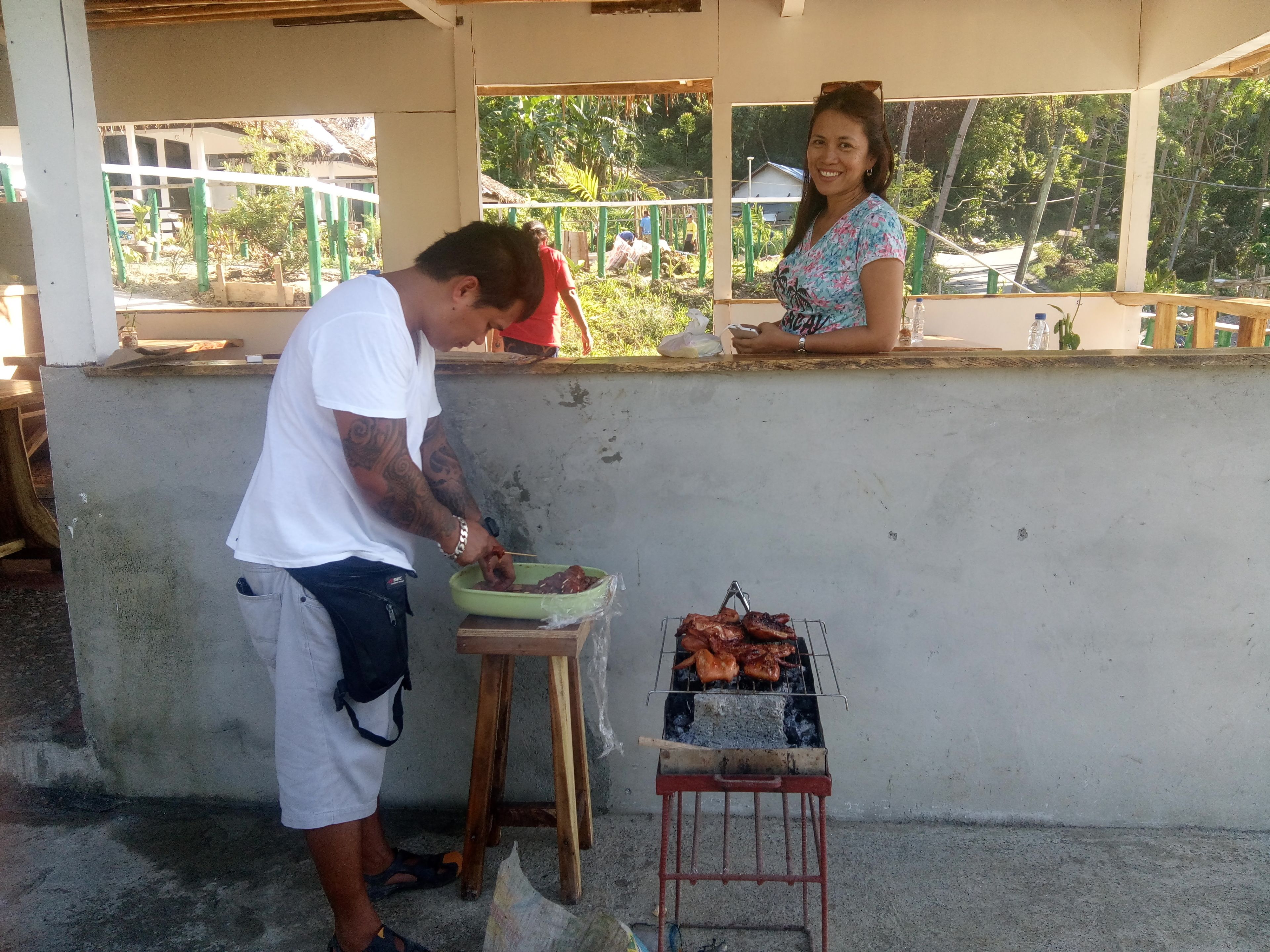 A man grilling meat on a portable grill outside a building with a woman standing nearby.