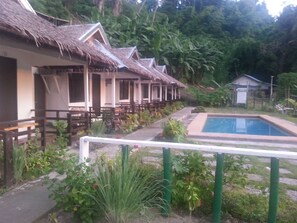 A row of thatched-roof cottages with outdoor seating, a swimming pool, and lush greenery. - Dahilig Resort (Puerto Galera)