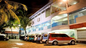 Un edificio de varios pisos con estacionamiento, palmeras y fachada bien iluminada.