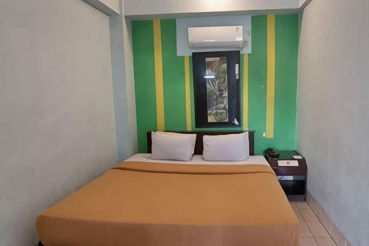 Una cama individual con una colcha marrón, una mesita de noche con un teléfono y una ventana con vistas a la vegetación.