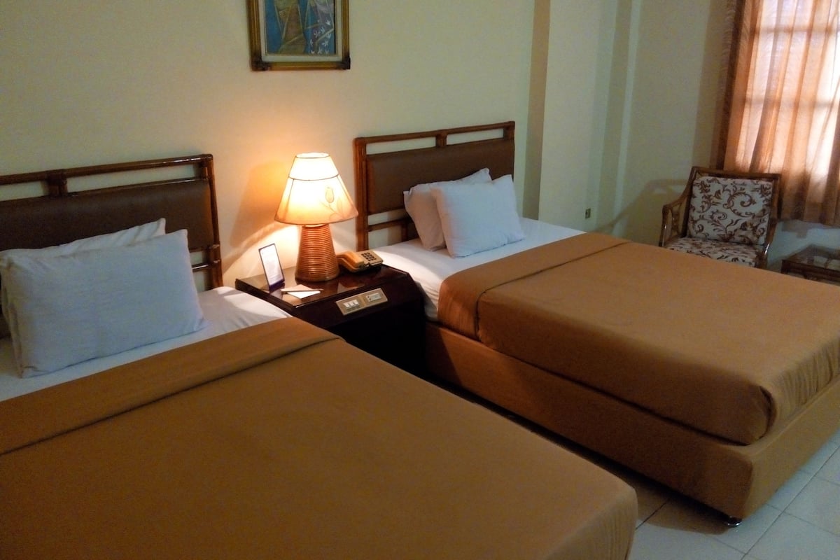 Habitación de hotel con dos camas, una mesita de noche con una lámpara, una silla y un cuadro en la pared.