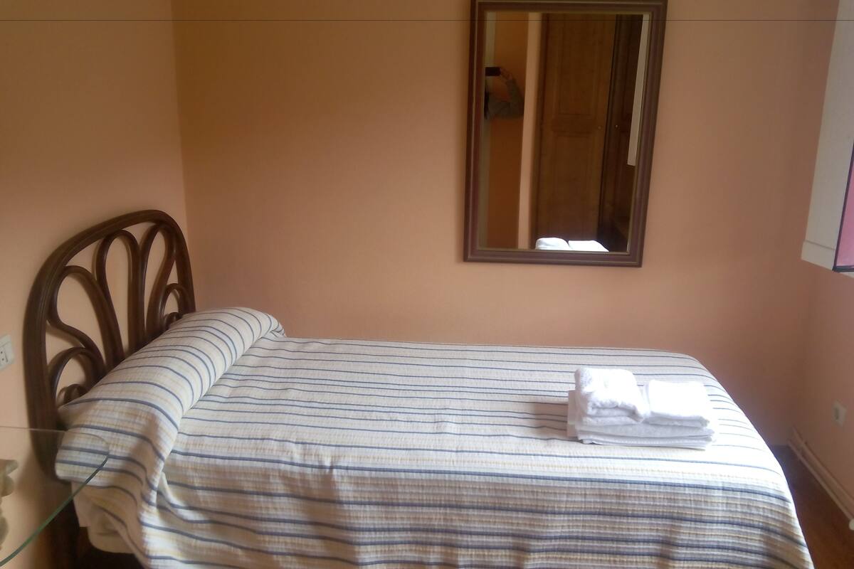 Una habitación pequeña con cama individual, cabecera de madera, cubrecama a rayas y una toalla doblada con cuidado sobre la cama.
