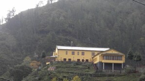 A yellow building with a flat roof, situated on a hillside with dense forest in the background.