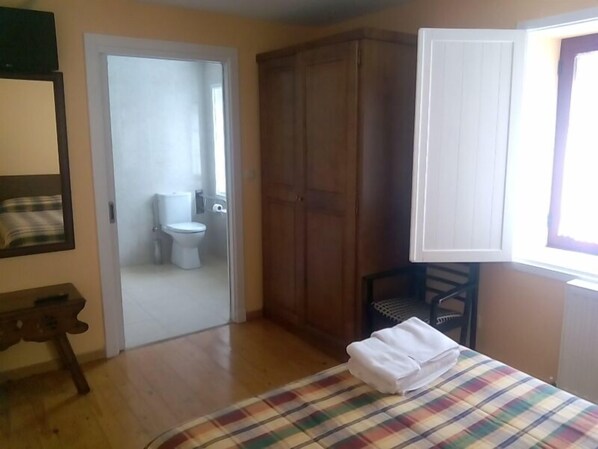 Une chambre à coucher avec un lit, une armoire en bois, une salle de bain visible à travers une porte ouverte et une fenêtre avec des rideaux.