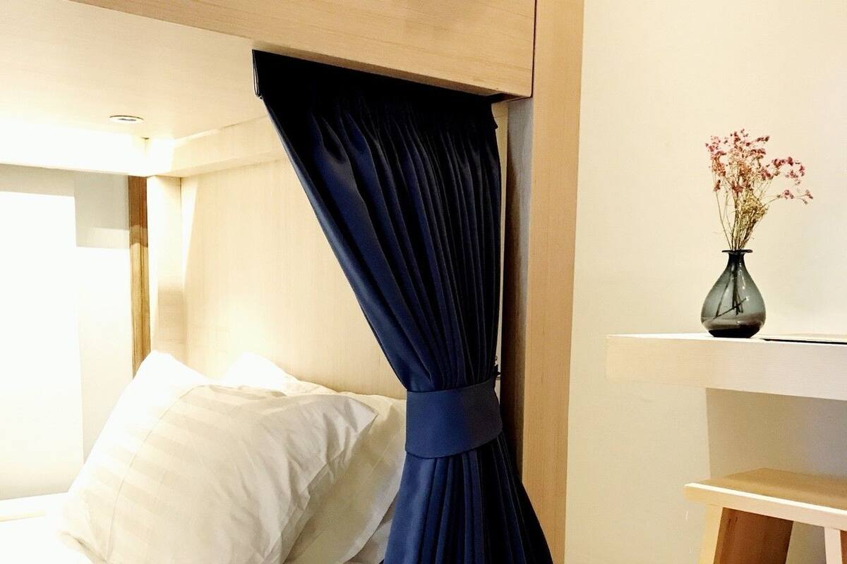 A bed with white linens, a dark blue curtain, and a small wooden nightstand with a vase of dried flowers.
