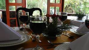 A dining table set with white plates, silverware, and wine glasses, with a view of greenery through large windows. - Eeescart Family Resort (Bandarawela)