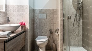 Un baño moderno con tocador de madera, lavamanos blanco, inodoro y zona de ducha con mampara de vidrio.