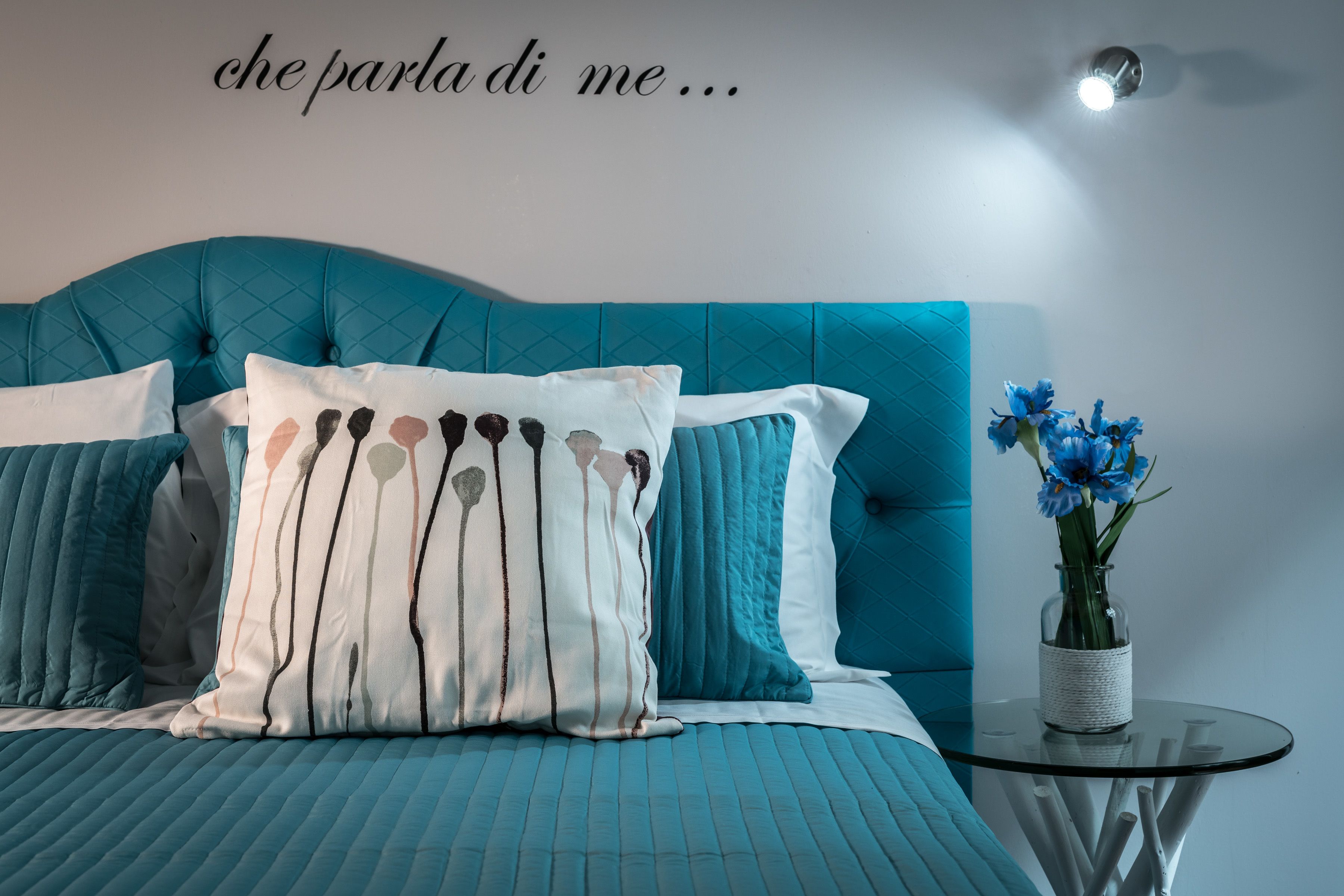 Une chambre à coucher avec une tête de lit recouverte d’un tissu turquoise, un oreiller blanc orné d’un motif noir et blanc, et un vase rempli de fleurs bleues posé sur une table en verre.