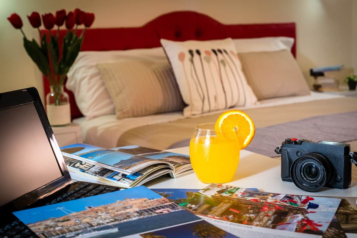 A laptop, a camera, a glass of orange juice, and a magazine on a bed with a red headboard.