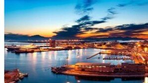 Un paisaje urbano costero al atardecer, con un puerto, edificios frente al agua y un puente destacado.