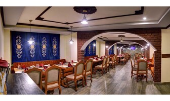 Un restaurante con mesas y sillas de madera, cortinas azules con diseños florales y paredes de ladrillo.