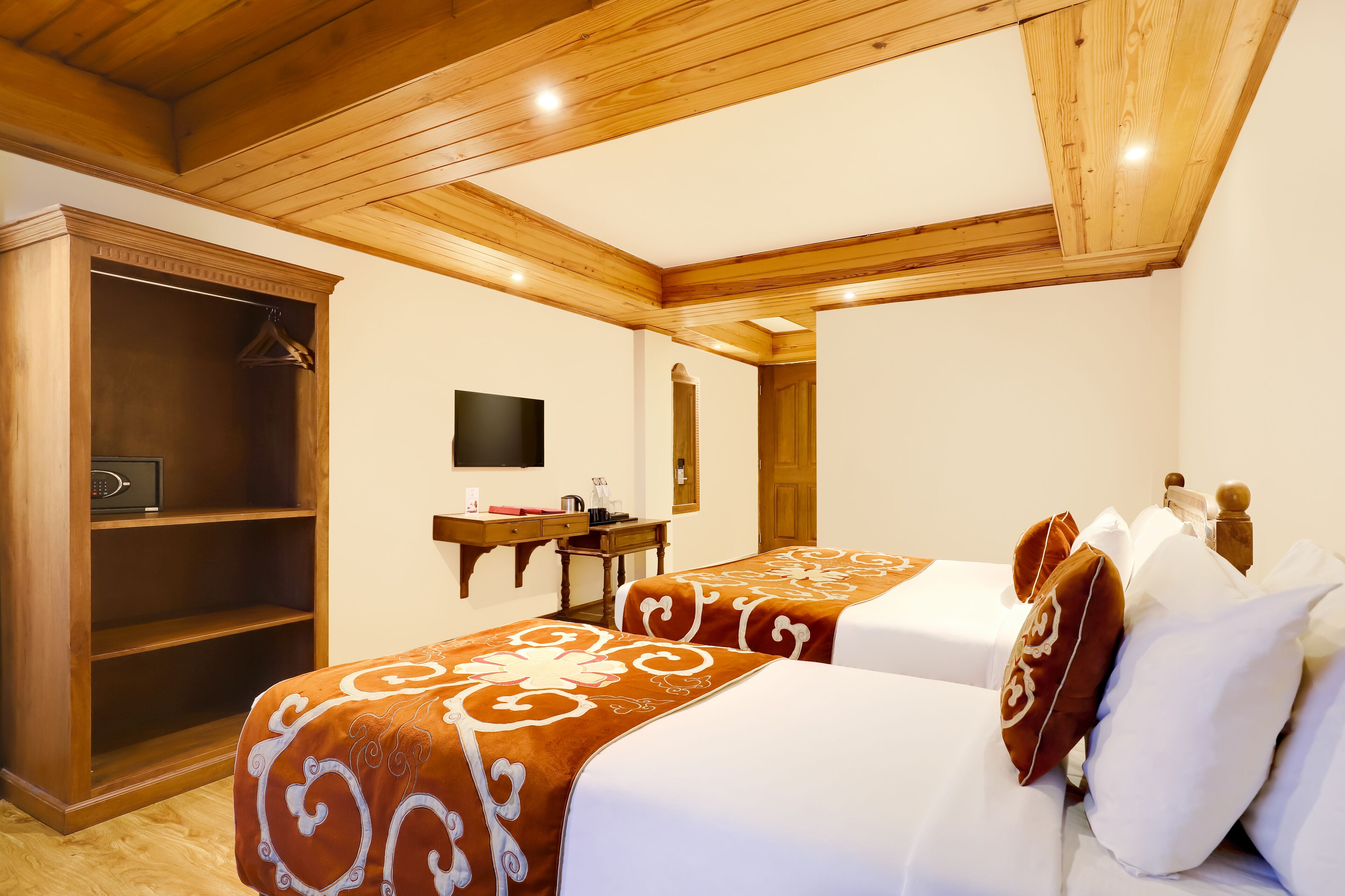 A hotel room with two beds, a wooden wardrobe, a desk with a flat-screen TV, and a wooden ceiling.