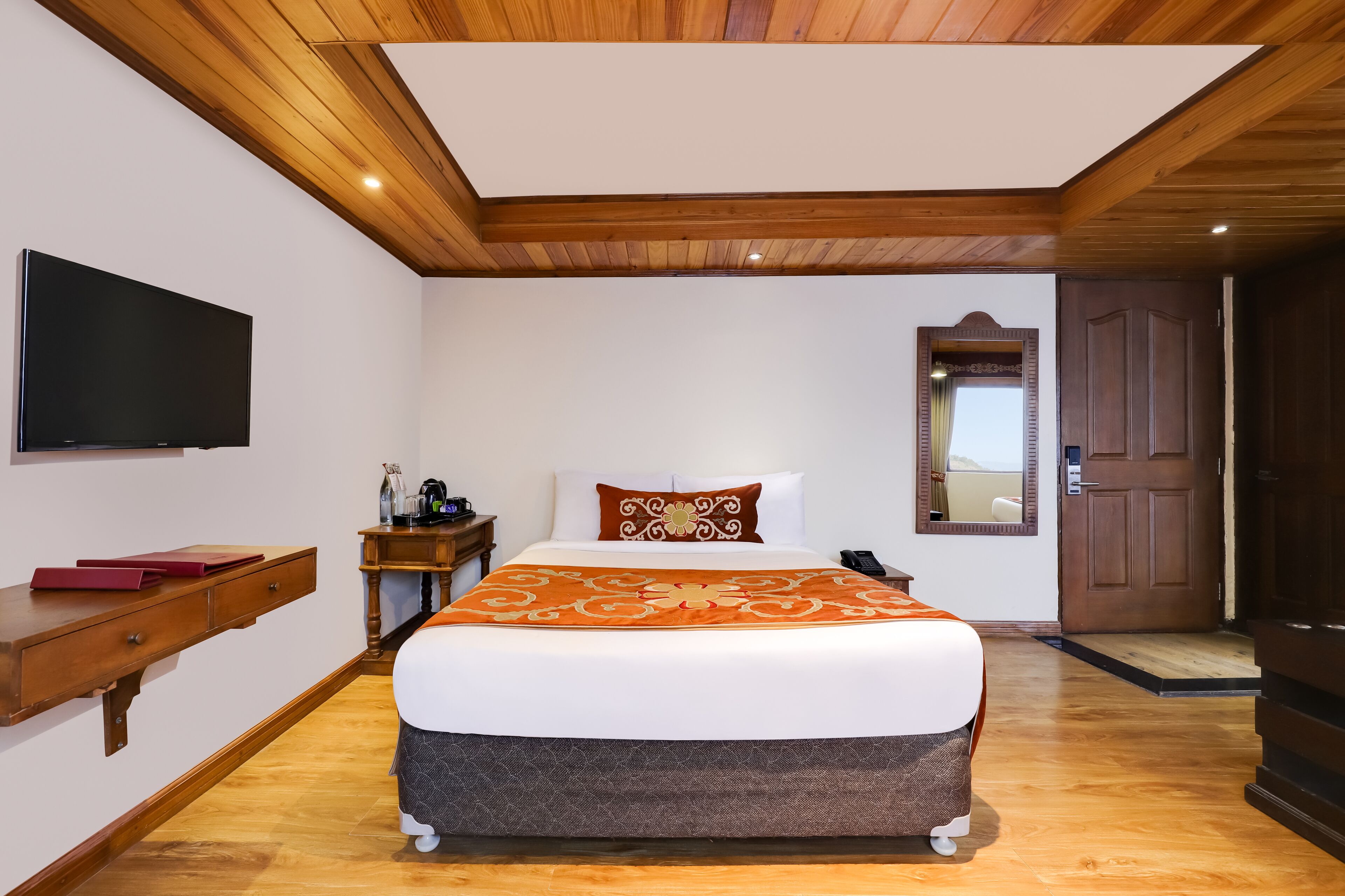 A hotel room with a wooden ceiling, a large bed with a patterned bedspread, a flat-screen TV mounted on the wall, a bedside table with a lamp, and a door leading to another room.