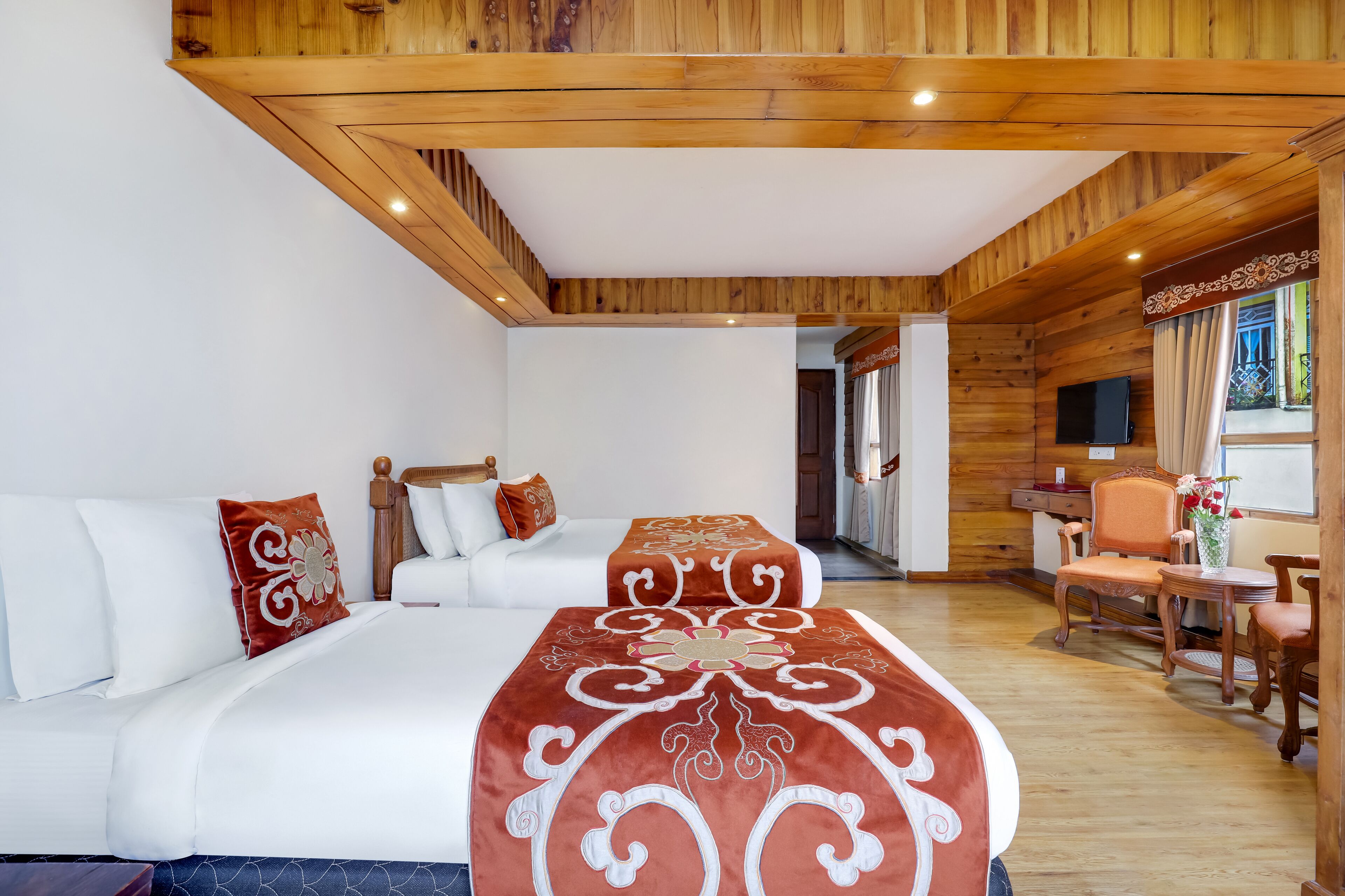 A hotel room with a wooden ceiling, a bed with white bedding, a sofa with white and red pillows, a small table with a vase, and a TV on the wall.