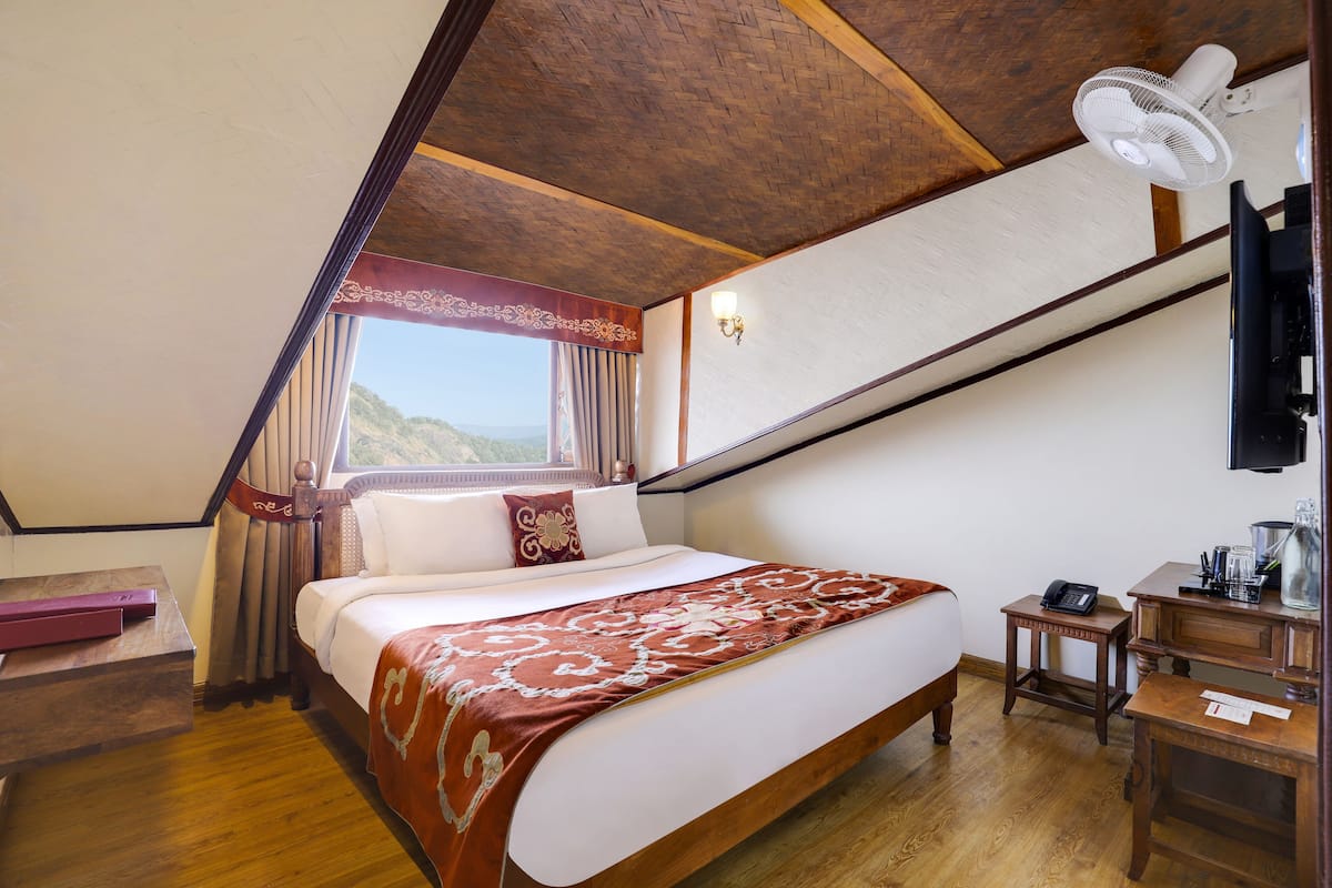 Une chambre avec un plafond en bois, un lit recouvert d’une couvre-lit à motifs, une table de chevet, une commode, un bureau, une chaise, une lampe, une fenêtre avec des rideaux et un ventilateur fixé au mur.