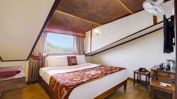 A bedroom with a wooden ceiling, a bed with a patterned bedspread, a bedside table, a nightstand, a desk, a chair, a lamp, a window with curtains, and a wall-mounted fan.