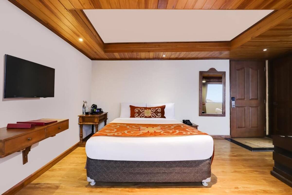 Une chambre avec un plafond en bois, un lit recouvert d’une couvre-lit à motifs, un téléviseur à écran plat, une table de chevet avec une lampe et une porte.