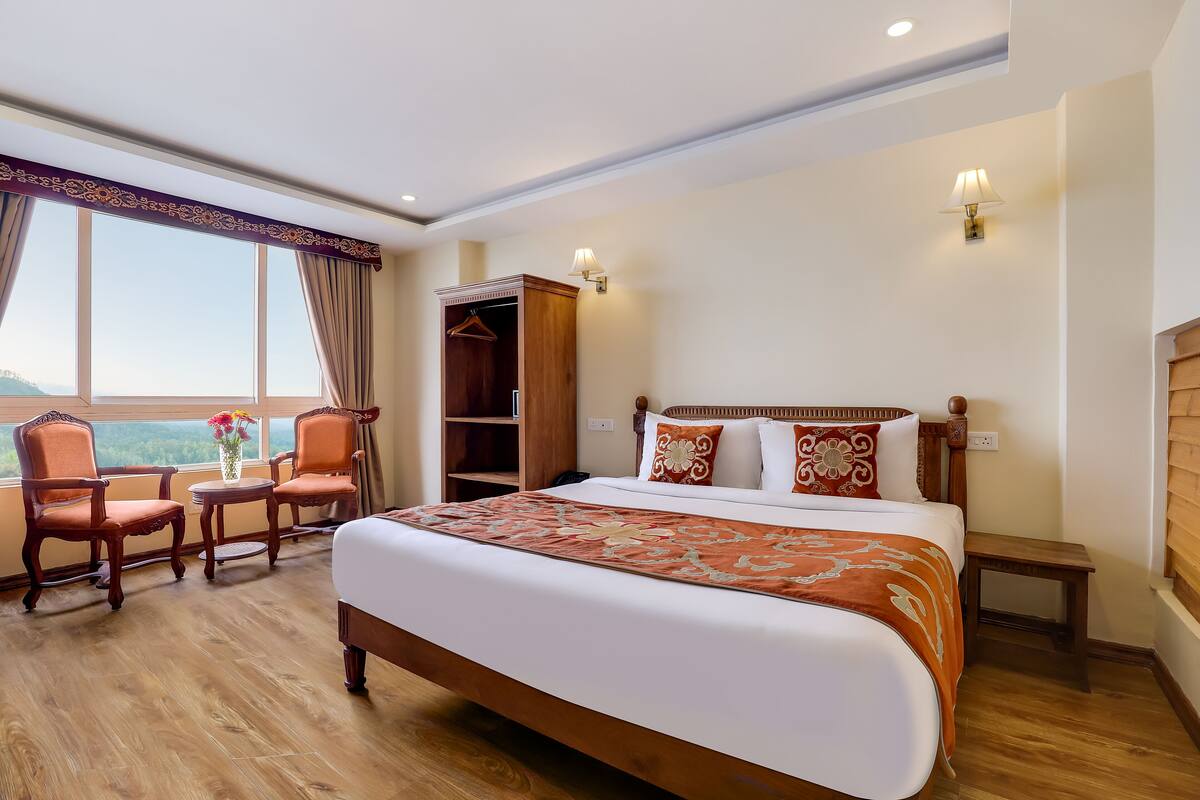 Une chambre d’hôtel avec un grand lit, deux chaises, une petite table, une armoire et une fenêtre donnant sur un beau paysage.