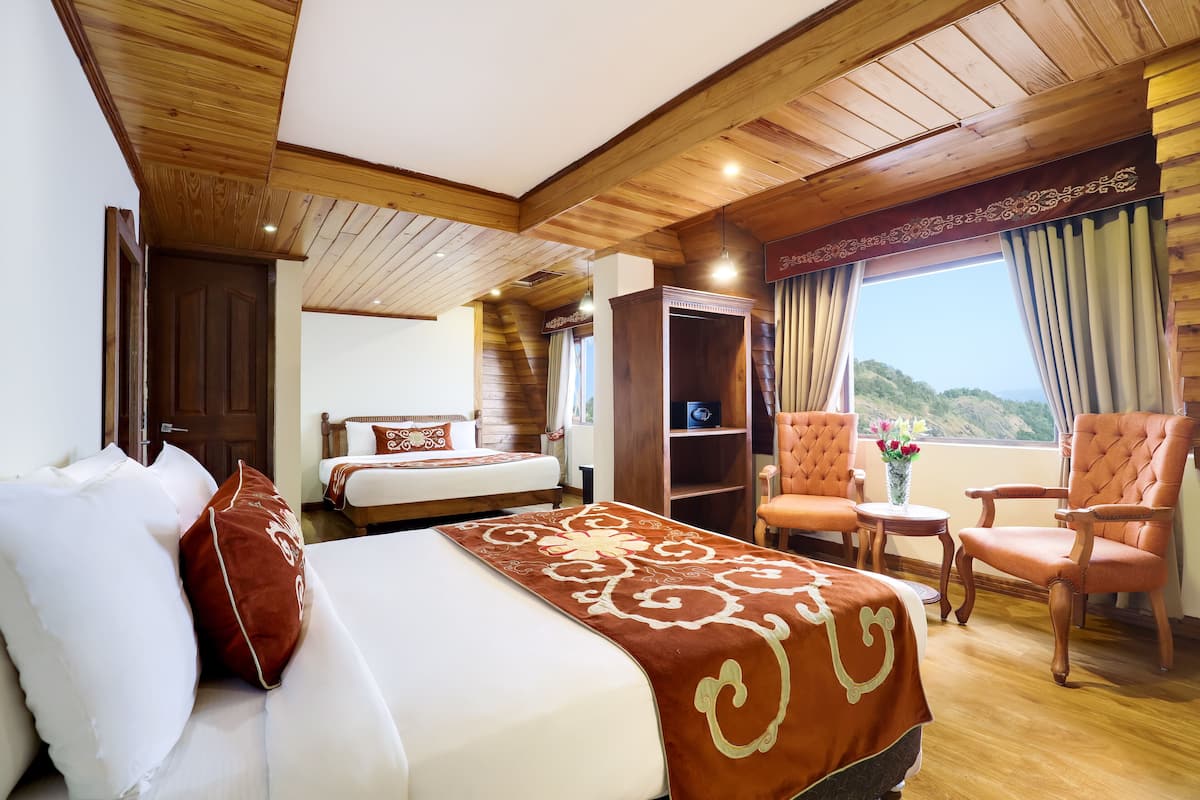 Une chambre d’hôtel avec un plafond en bois, un grand lit recouvert d’une courtepointe marron, un coin salon avec un fauteuil et une petite table, et une vue sur un paysage montagneux par la fenêtre.