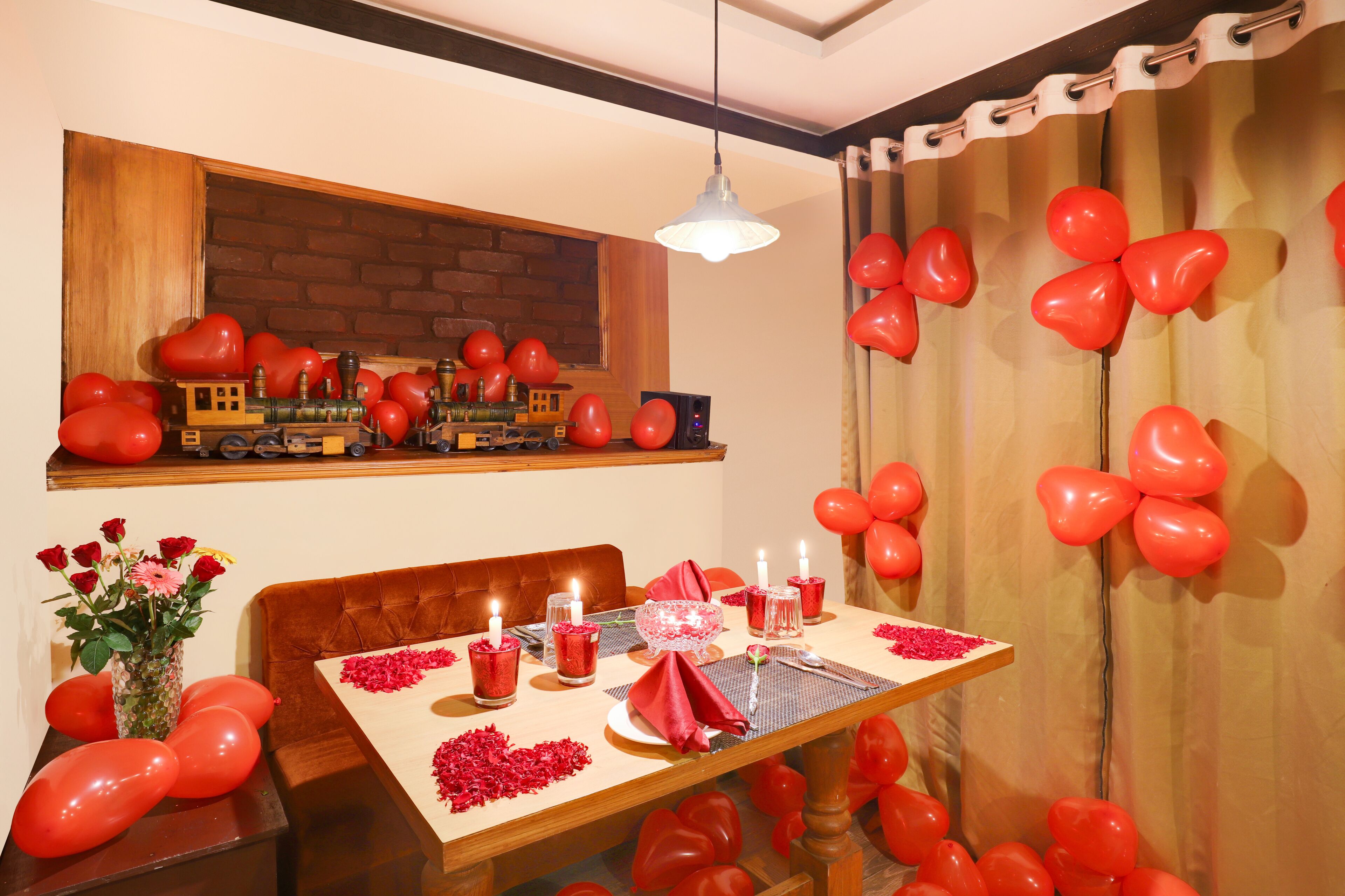 A dining area with a wooden table set for two, surrounded by red balloons and a toy train display on the wall.