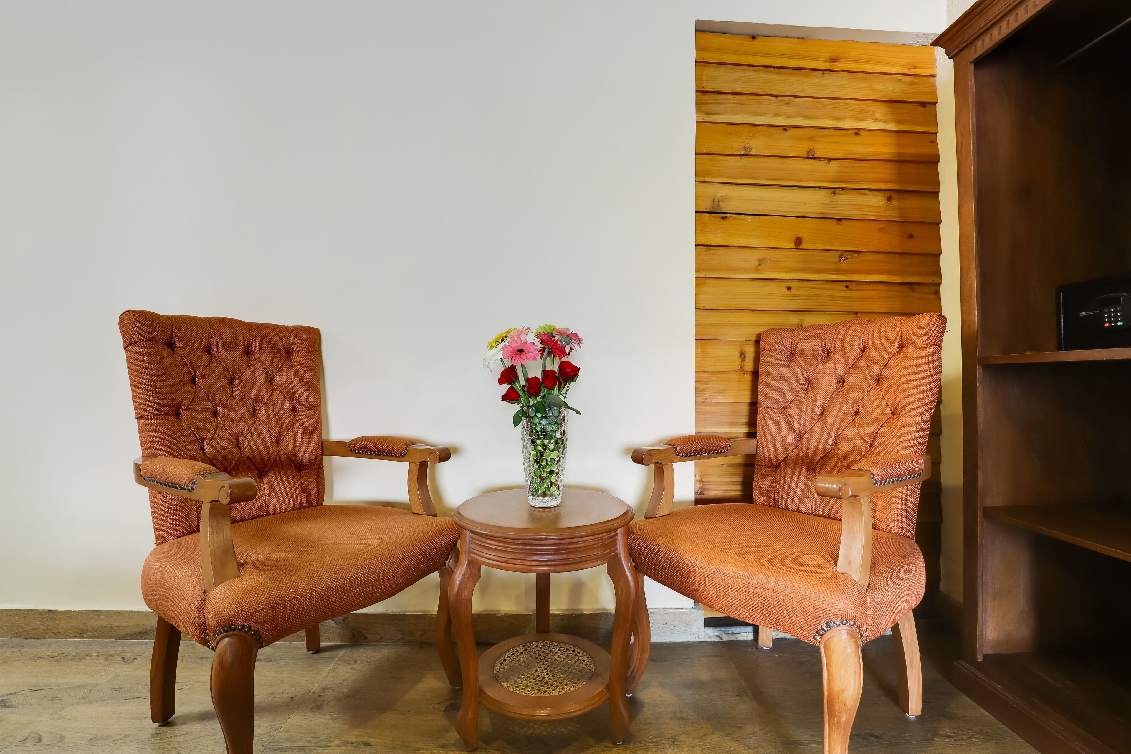 Dos sillones tapizados con reposabrazos y patas de madera, una mesita redonda de madera con un centro tejido, y un jarrón con flores sobre la mesa.