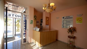 Un espace d’accueil d’hôtel avec un comptoir en bois, un lustre et divers affiches d’information accrochées aux murs.