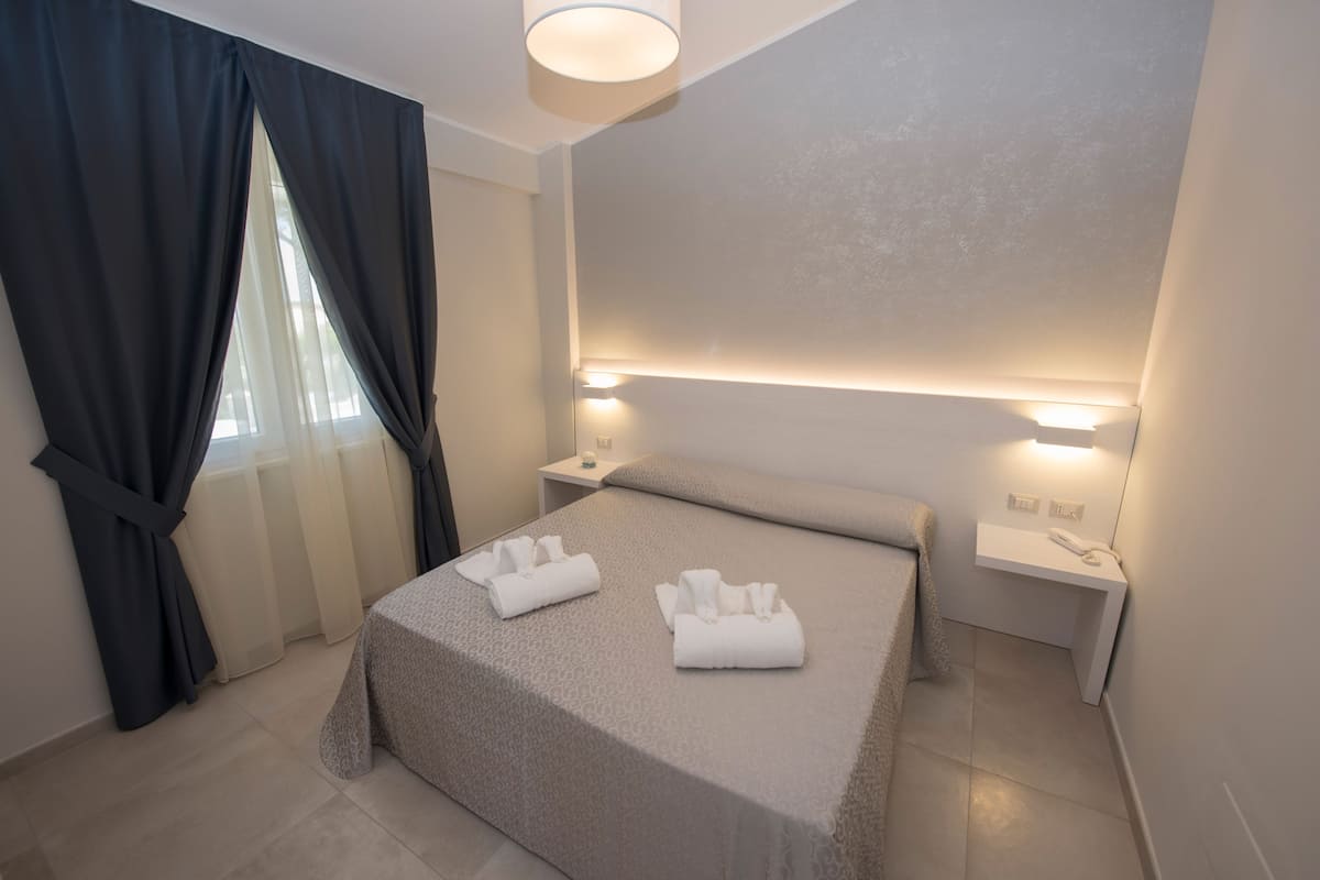 Una habitación de hotel con una cama bien hecha, dos mesitas de noche con lámparas y una ventana con cortinas.