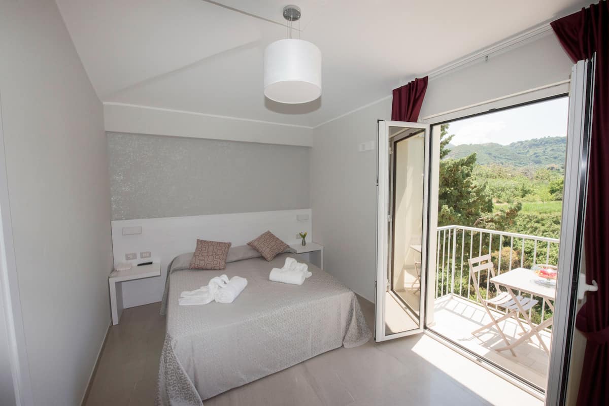 Un dormitorio con cama, mesita de noche, ventana con vista a la vegetación y balcón con mesa y sillas.