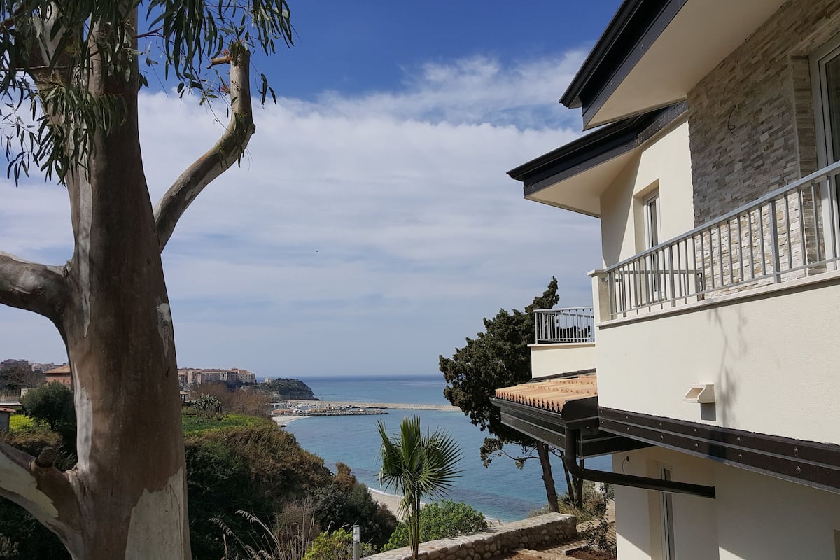 Un edificio costero con balcón que da al mar, un árbol grande en primer plano y cielo despejado.