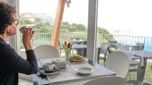 A person sitting at a table with a cup, a plate with food, and a vase with flowers. The table is set on a balcony with a view of the sea and mountains. - Borgo degli Dei - Affittacamere Poseidone (Parghelia)