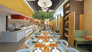 Ein modernes Restaurant mit einem langen Gemeinschaftstisch, separaten Sitzplätzen, einem zentralen Baum und einer linksseitig sichtbaren Küche.