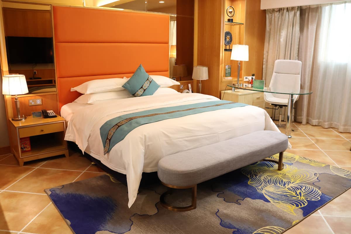A hotel room with a large bed, a glass table, a chair, a TV, and a bedside table with a lamp.