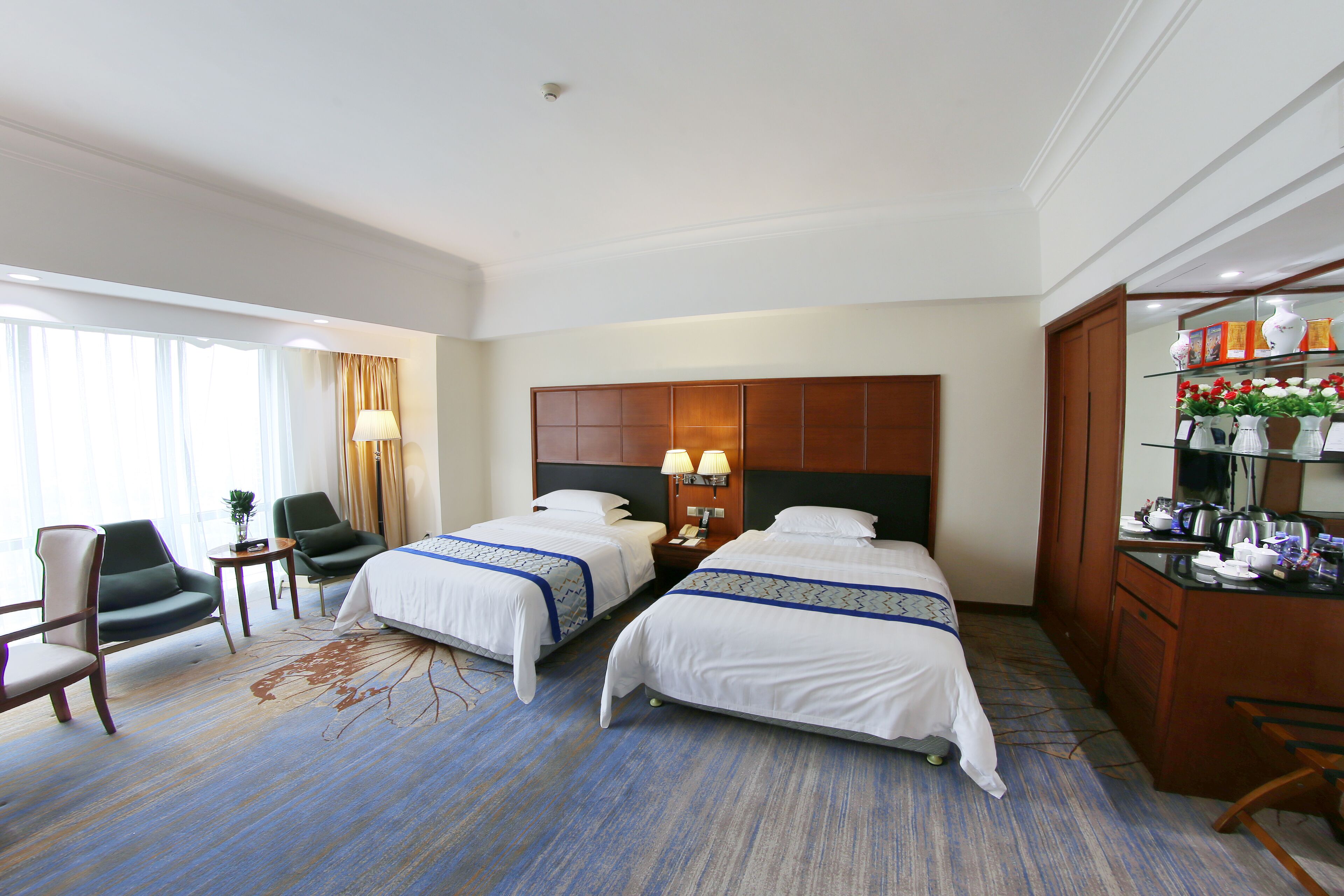 A hotel room with two beds, a desk, a chair, a lamp, and a view of the outdoors.