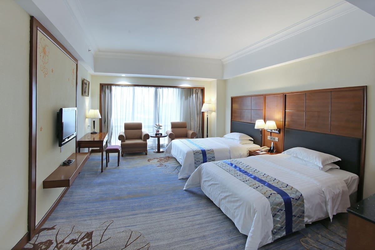 A hotel room with two beds, a TV, a desk, a chair, and a view of the outdoors through large windows.