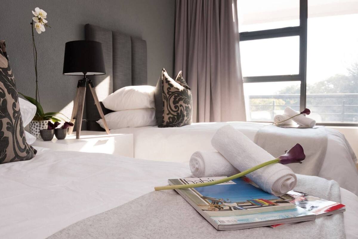 A hotel room with a bed, pillows, a lamp, a window, and a magazine on the bedside table.