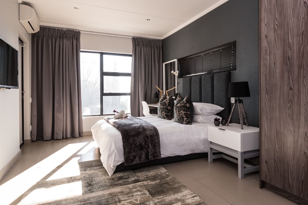 A modern bedroom with a large bed, a bedside table, a TV mounted on the wall, and a window with curtains.