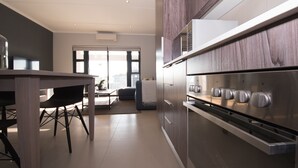 Een moderne keuken met een ingebouwde oven, een eethoek met een tafel en stoelen, en uitzicht op het buitenleven door een glazen deur.