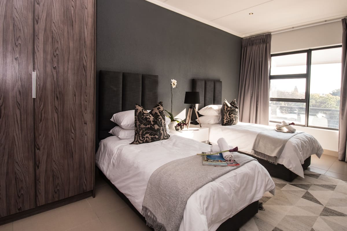 A modern hotel room with two beds, dark wood wardrobe, grey walls, large window with curtains, and a geometric patterned rug.