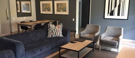 A modern living room with a dark blue sofa, two armchairs, a coffee table, and a dining area with a table and chairs.