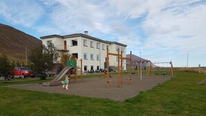 A playground with a slide and climbing structures, surrounded by a grassy area and a building in the background. - Húsabakki Guesthouse (Dalvik)