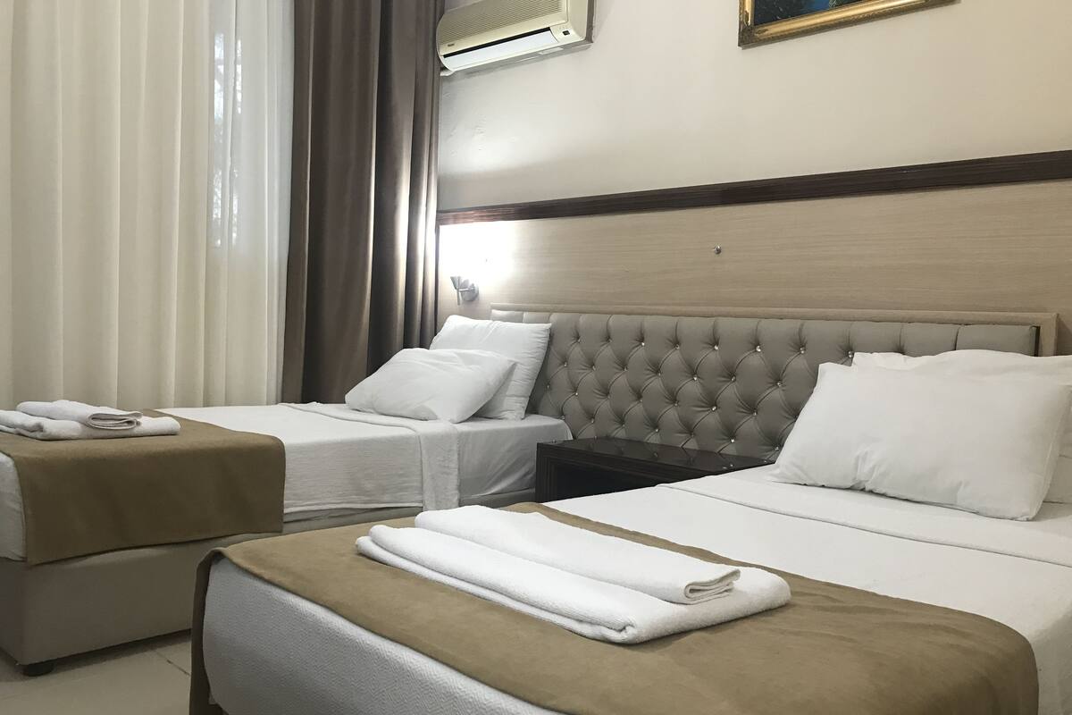 A hotel room with two beds, each with white linens and a brown throw blanket. There is a nightstand with a lamp between the beds, and an air conditioning unit mounted on the wall.