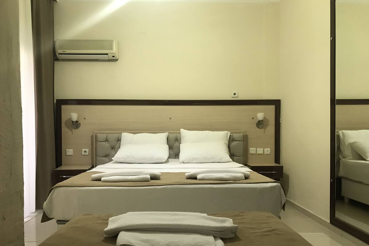 A hotel room with a bed, a bedside table, a nightstand, an air conditioning unit, and a mirror.