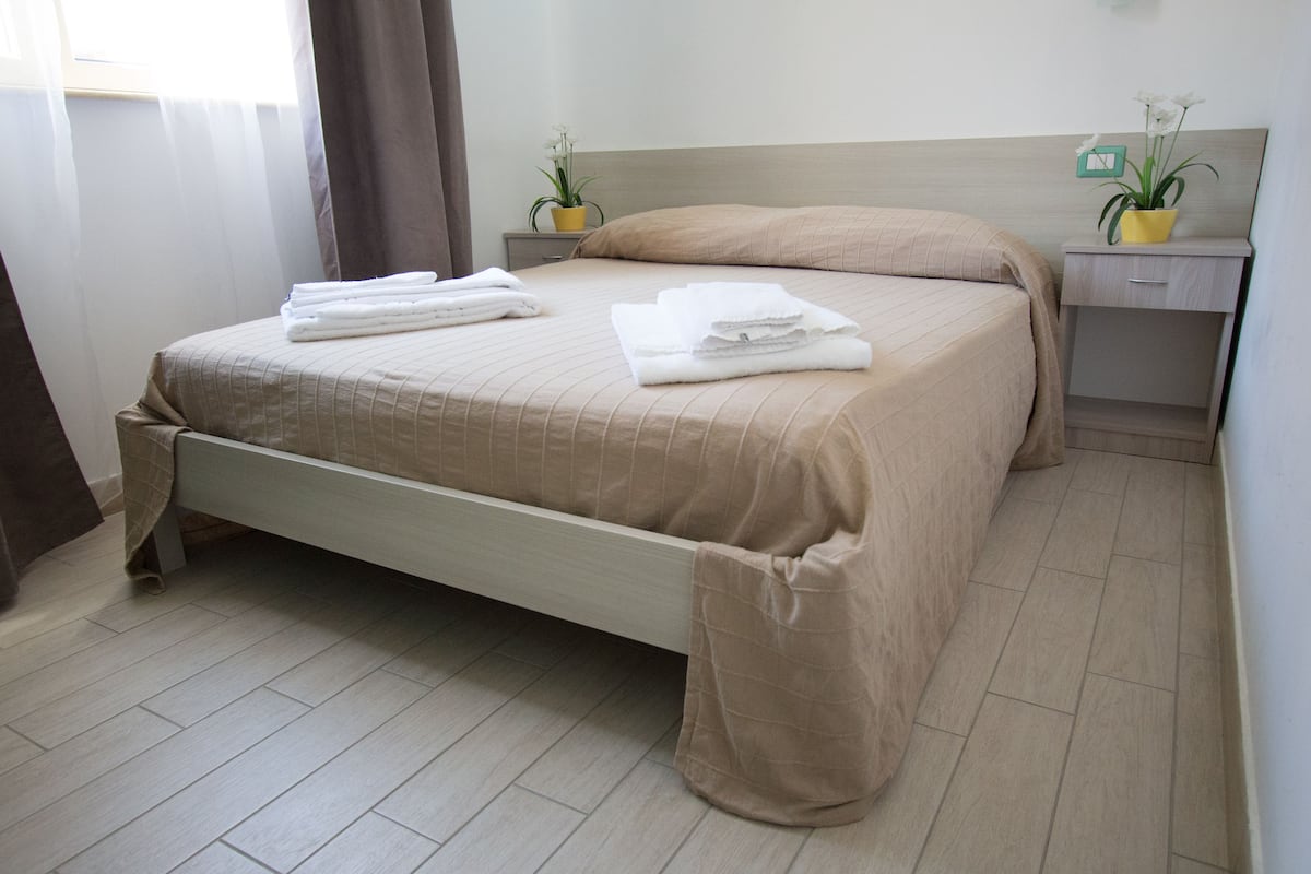 A neatly made bed with a beige bedspread and pillows, a bedside table with a plant, and a nightstand with a clock.