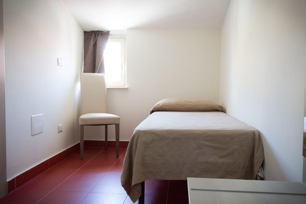 A small, sparsely furnished room with a single bed, a chair, and a window.