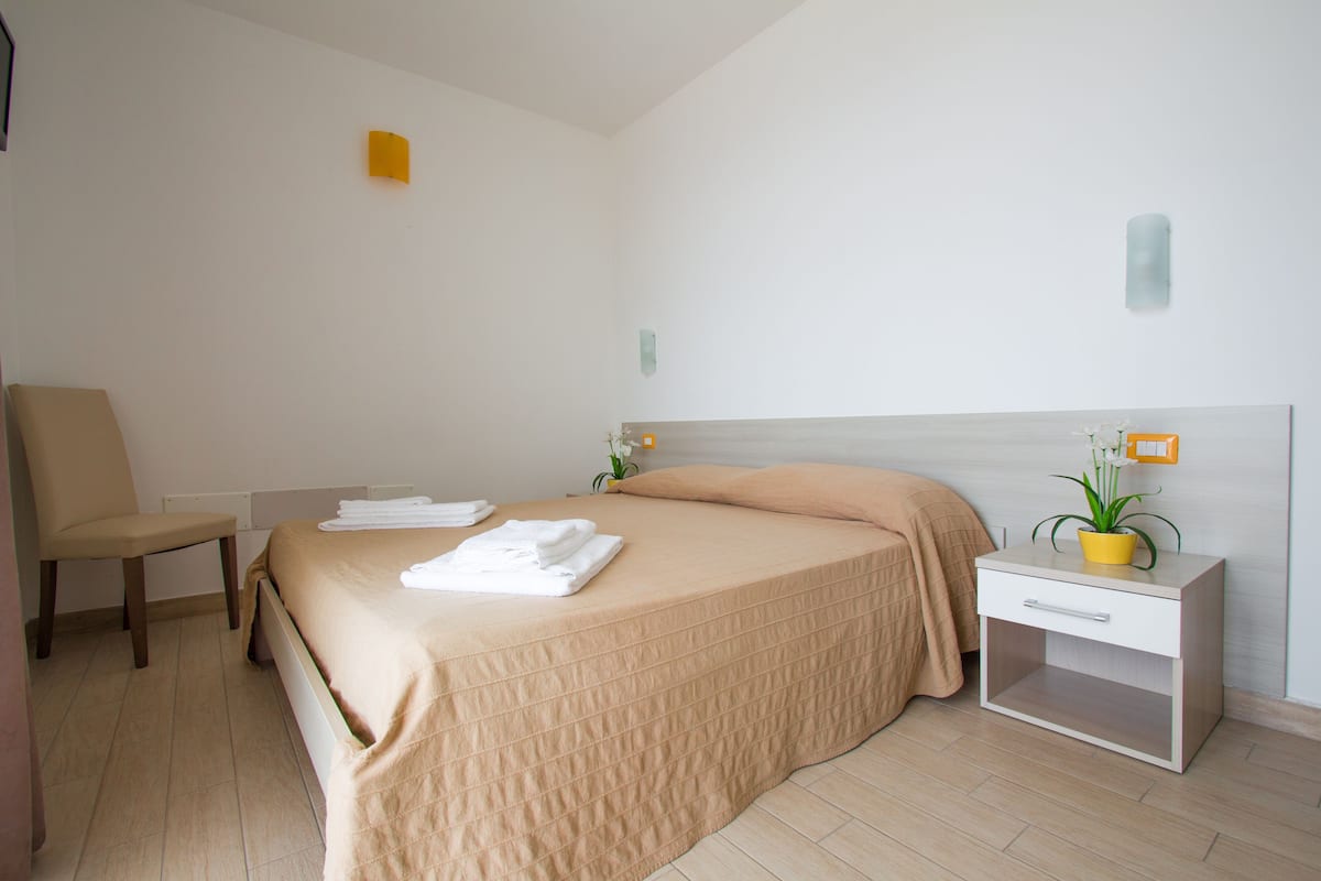 A hotel room with a bed, a chair, a nightstand with a plant, and a wall-mounted light fixture.