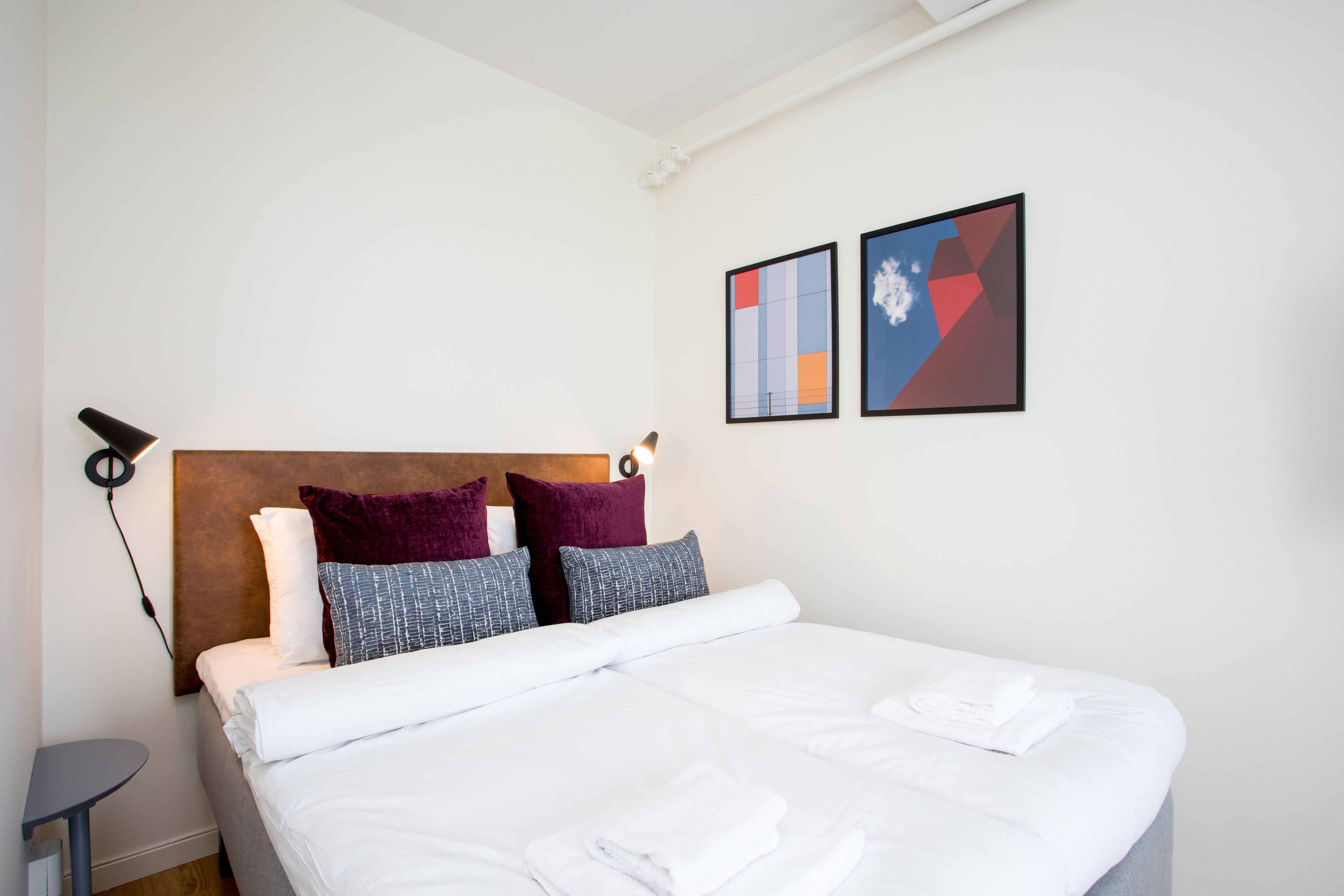 Een net opgemaakt bed met witte lakens en opgevouwen handdoeken, een rond nachtkastje en twee abstracte kunstwerken aan de muur.