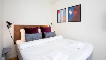 Een net opgemaakt bed met witte lakens en opgevouwen handdoeken, een rond nachtkastje en twee abstracte kunstwerken aan de muur.