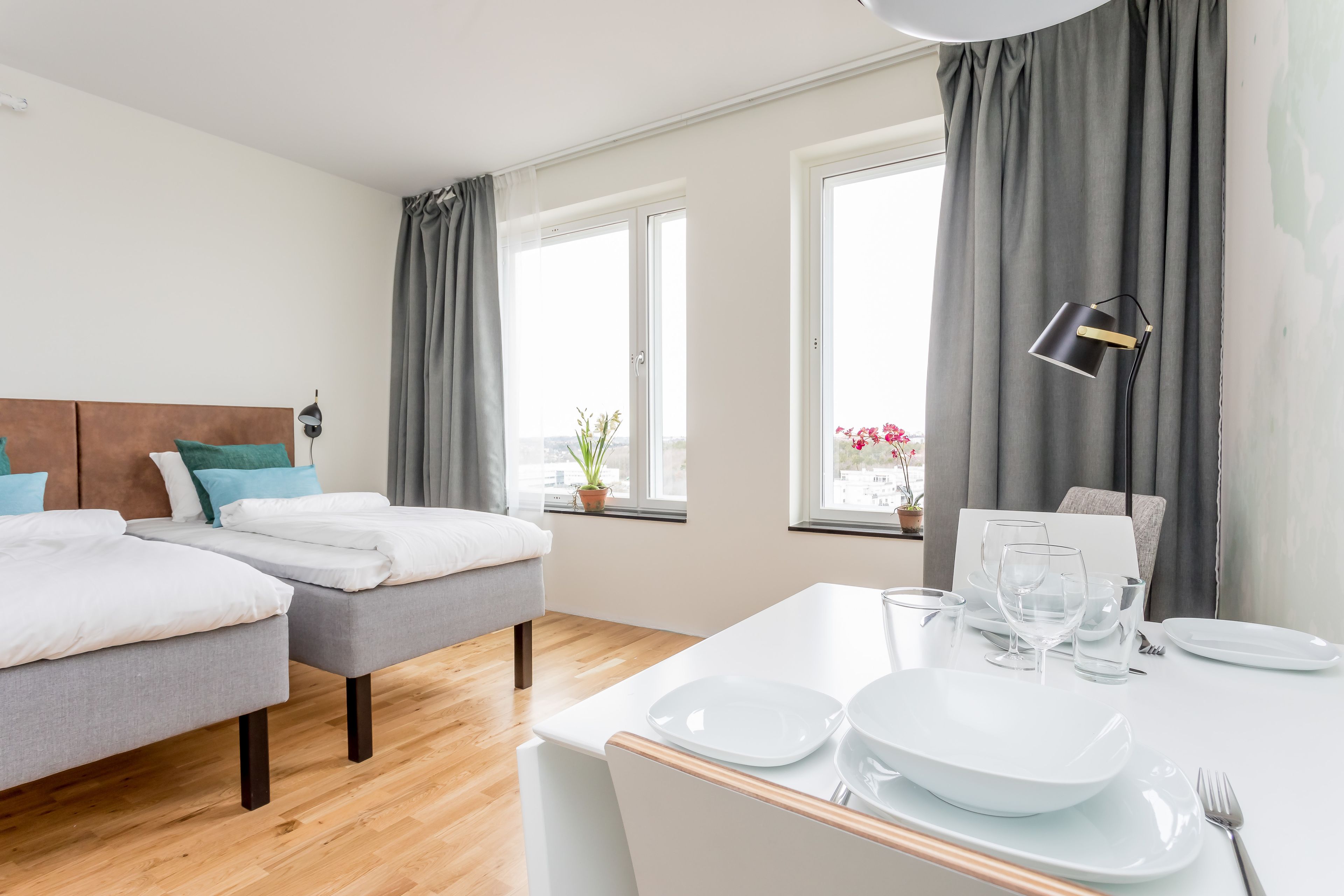 Une chambre d’hôtel avec deux lits, une table à manger dressée avec des assiettes et des verres, et une vue sur l’extérieur grâce à de grandes fenêtres.